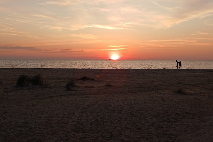 Two people walking on a beach at sunset with a calm sea and colorful sky.